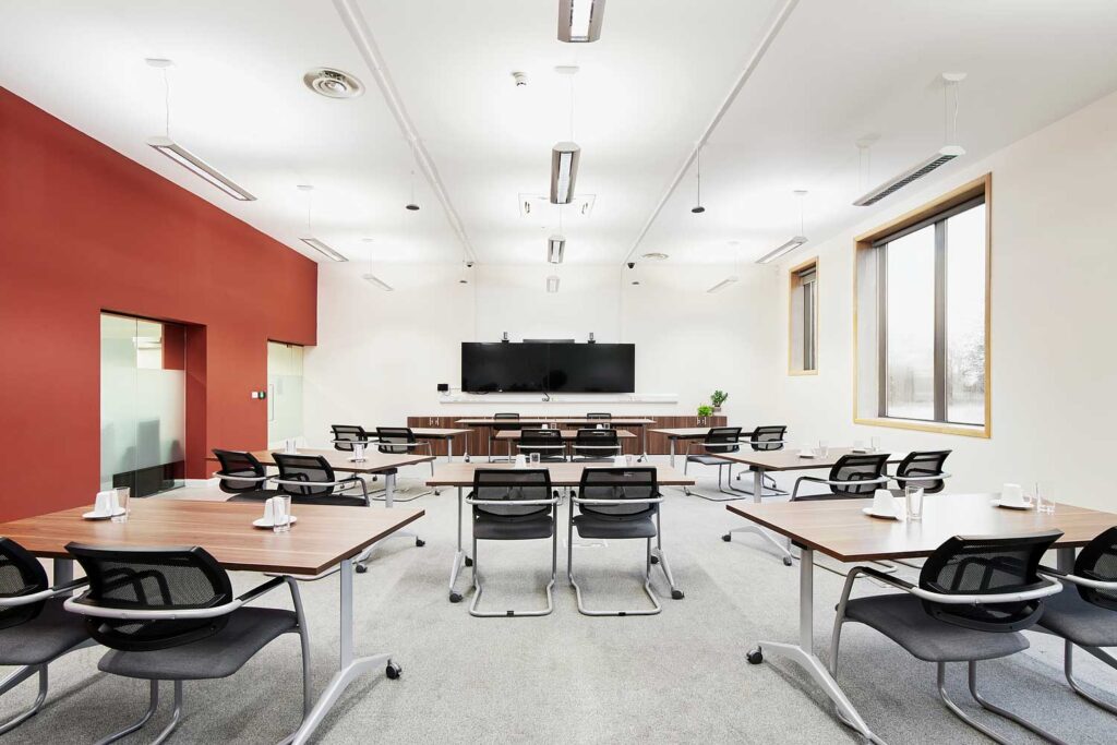 A large room with tables and chairs all facing towards a large screen at one end of the room.