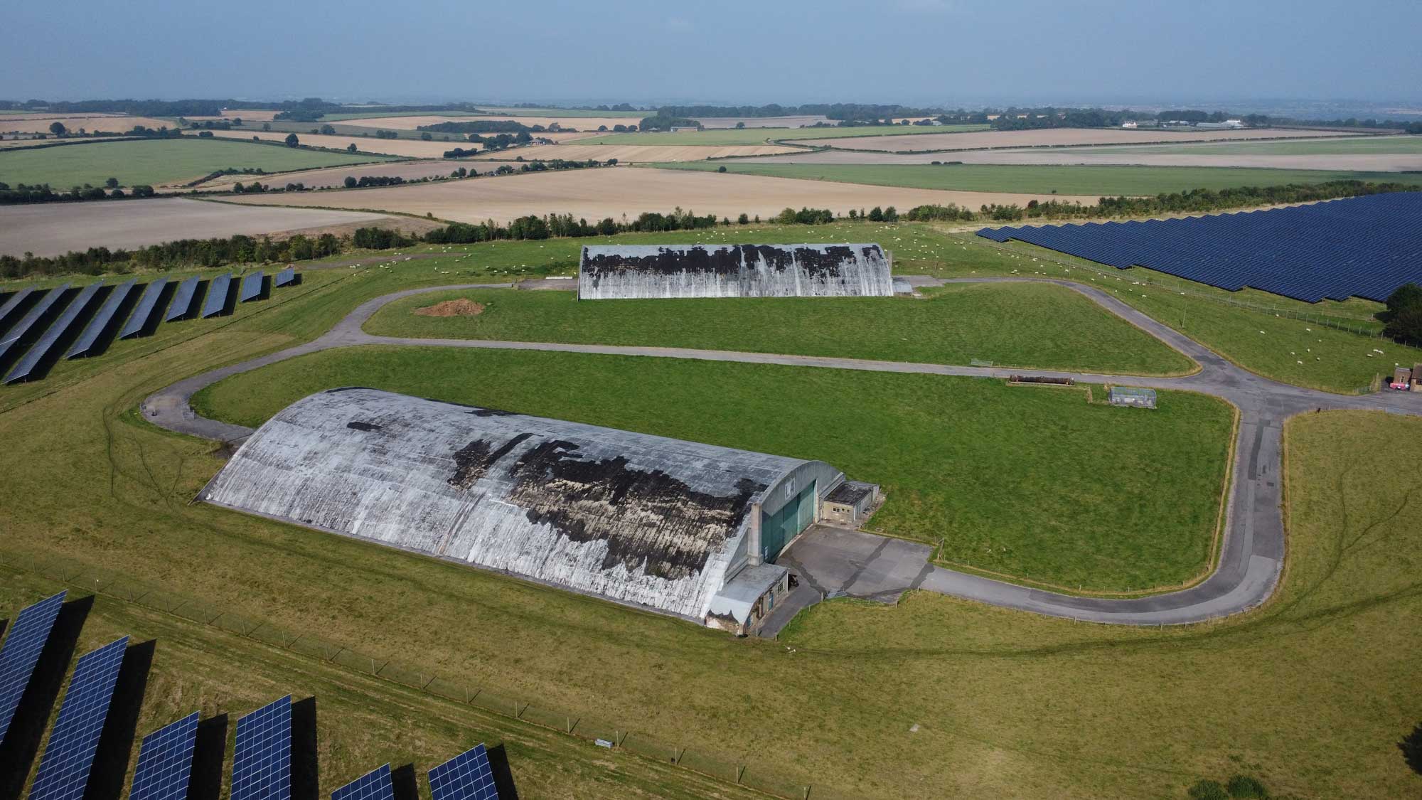 An aerial view of two old aircraft hangars and lots of solar panels in the English countryside.